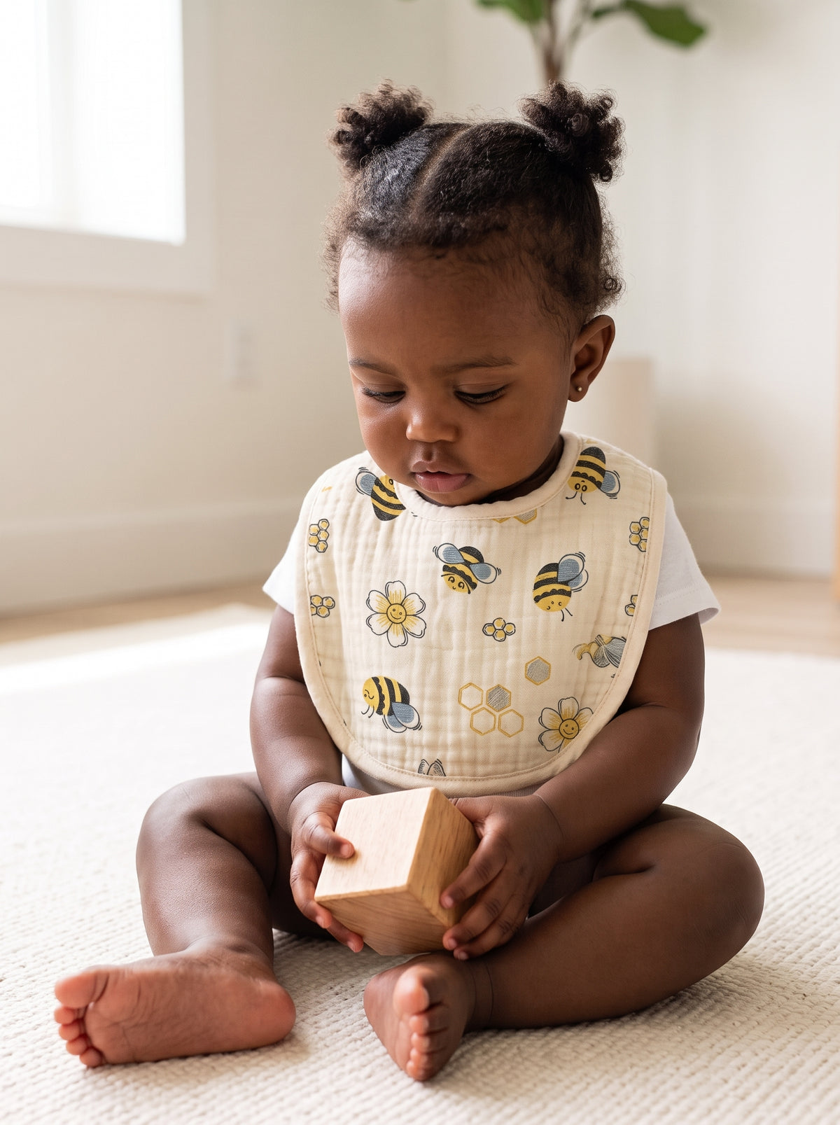 A nine-month-old baby girl sitting up on a cream rug wearing a cream mulmul muslin bib with a bee and daisy print, holding a wooden block. AI-generated image.