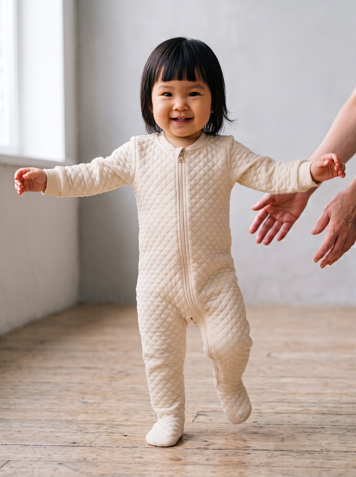An eighteen-month-old girl mid-step toward the camera in a cream quilted footed cotton sleepsuit, arms out for balance, an adult's open hands just behind her. AI-generated image.