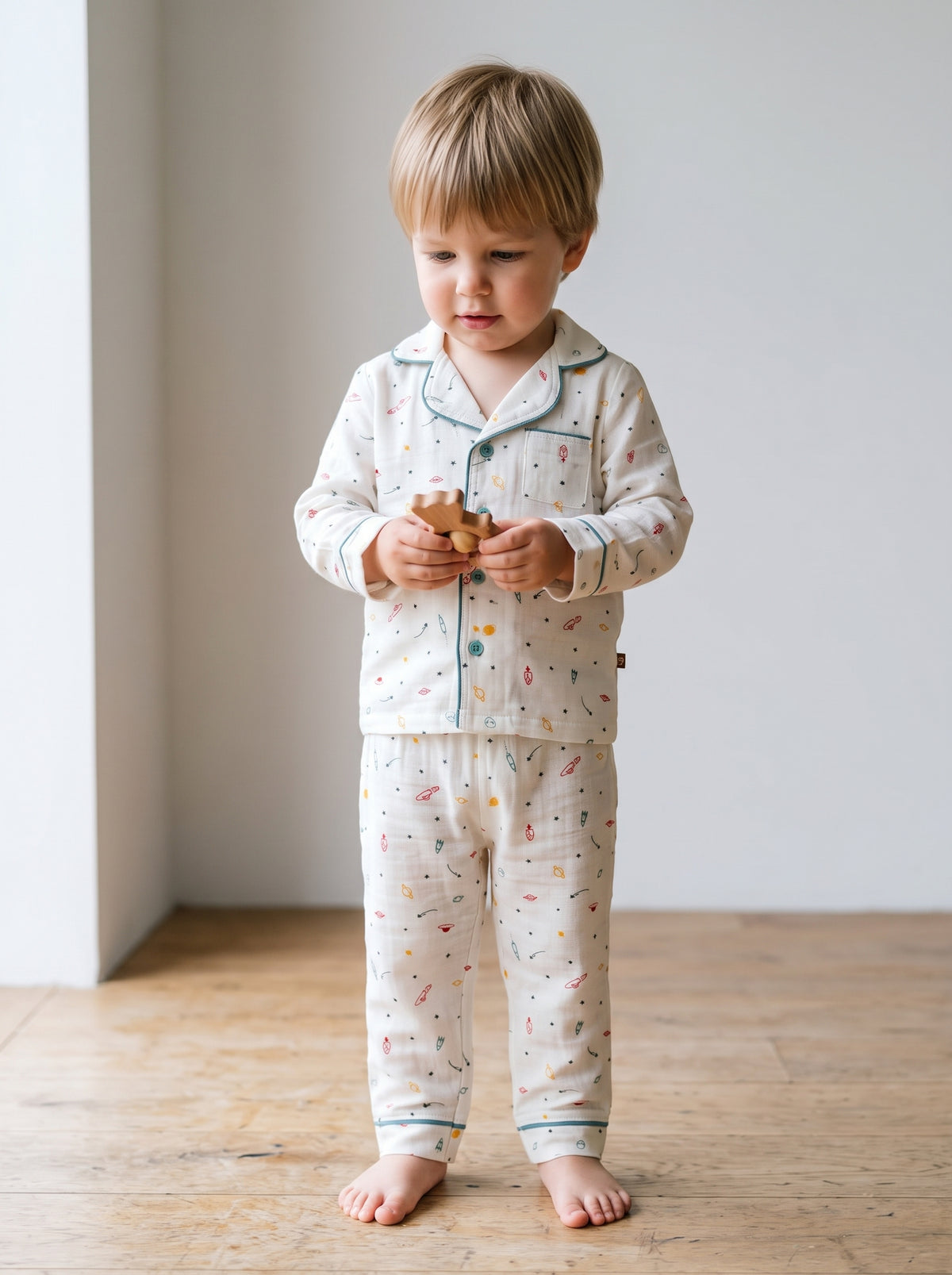 A two-year-old boy standing on a wooden floor in a cream mulmul muslin two-piece set with a collar and teal piping, looking down at a small wooden toy. AI-generated image.