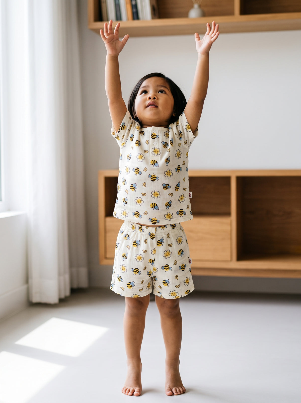 A two-year-old girl up on tiptoes with both arms stretched high, wearing a cream mulmul muslin two-piece set with a bee print. AI-generated image.