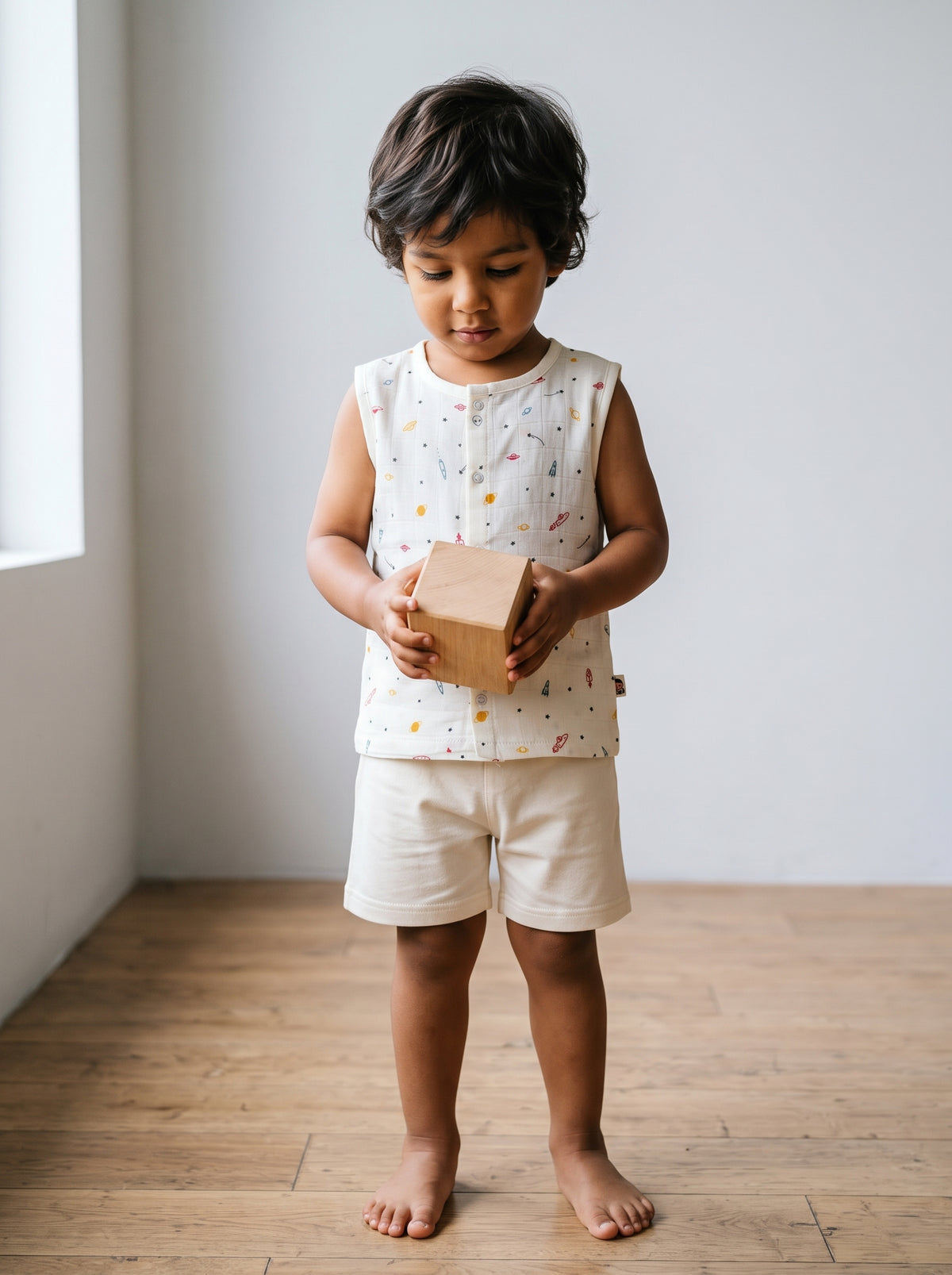 A three-year-old boy standing on a wooden floor in a cream mulmul muslin sleeveless top and cream shorts, holding a wooden block. AI-generated image.