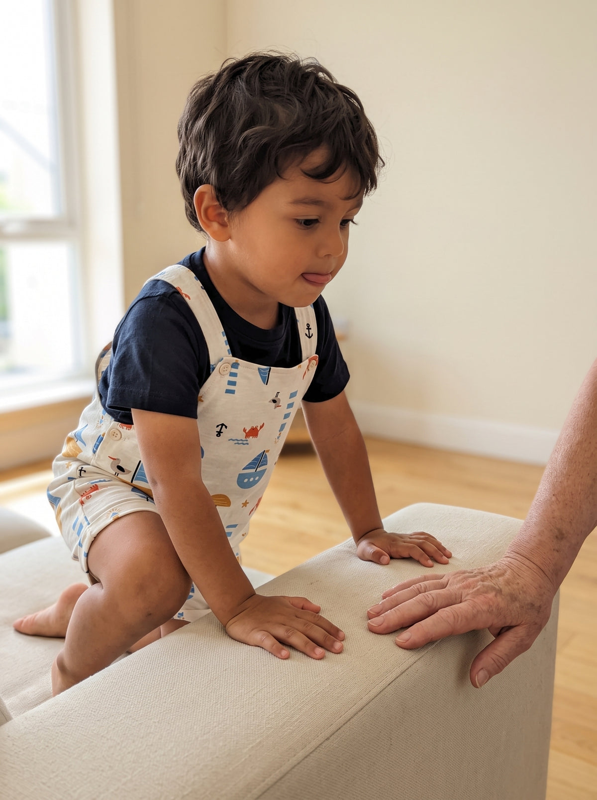 A three-year-old boy climbing onto a low cream sofa in cream cotton nautical-print overalls and a navy t-shirt, an older adult's hand resting on the cushion beside him. AI-generated image.