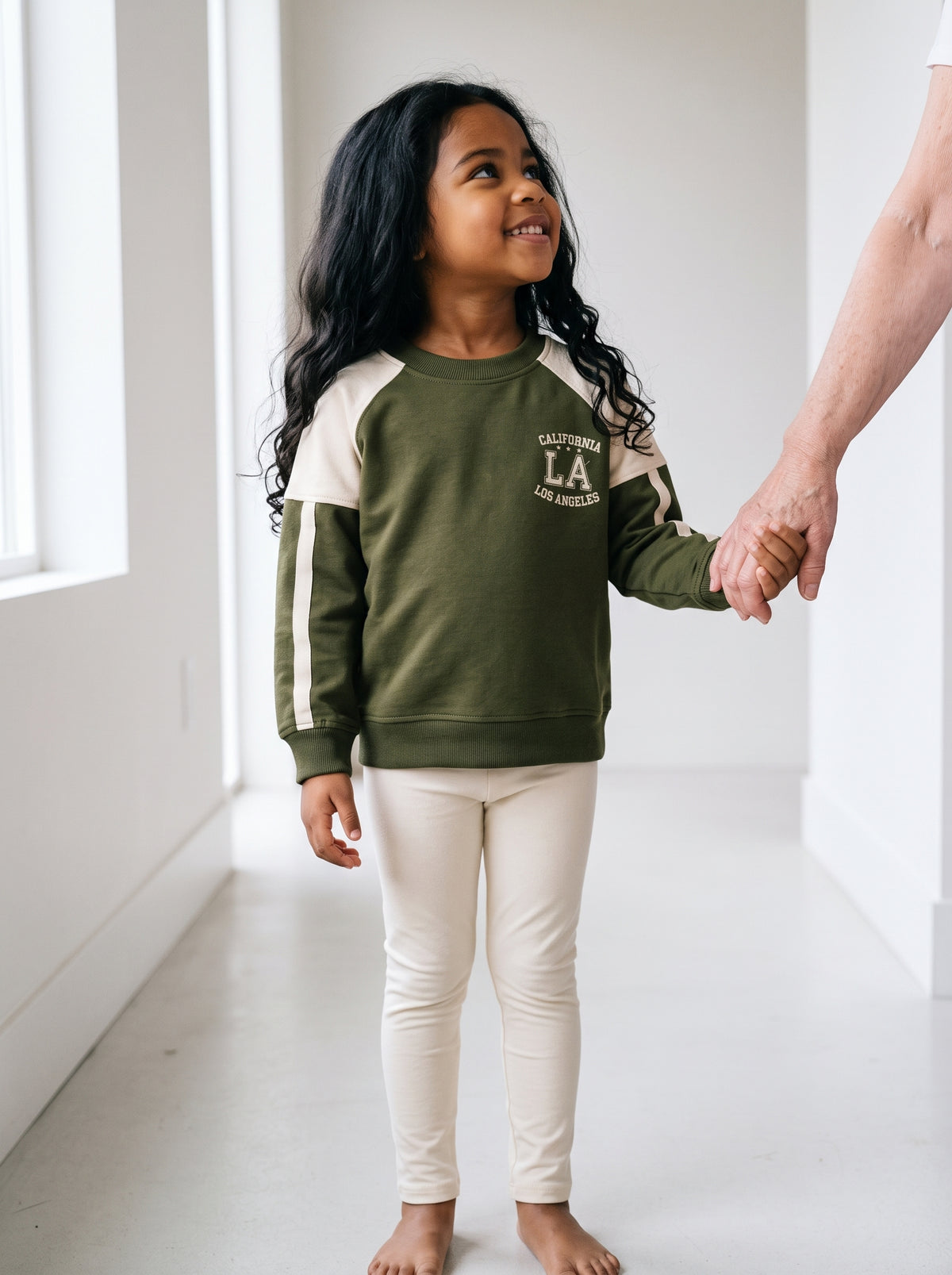 A five-year-old girl in an olive green cotton raglan sweatshirt holding an older adult's hand, looking up at them. AI-generated image.