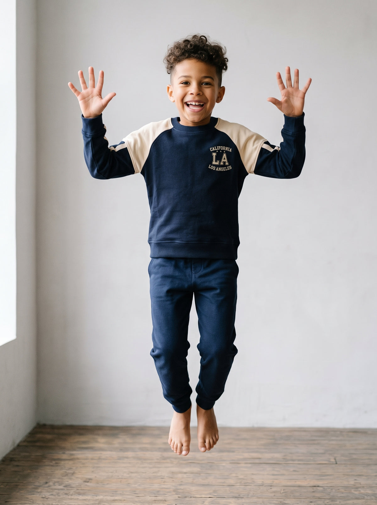 An eight-year-old boy caught mid-jump with both feet off the floor and both arms raised, in a navy cotton raglan sweatshirt with cream sleeve panels. AI-generated image.