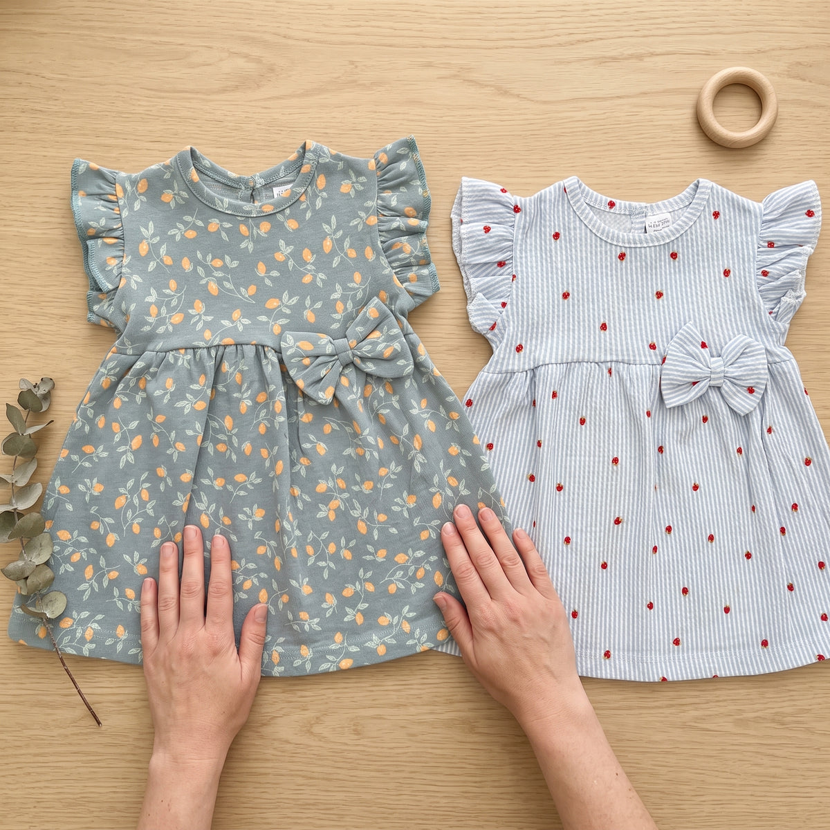 Two cotton dresses laid flat on a pale oak table, hands smoothing the skirt of the blue-grey dress with the orange print, the blue and white striped strawberry dress beside it. AI-generated image.