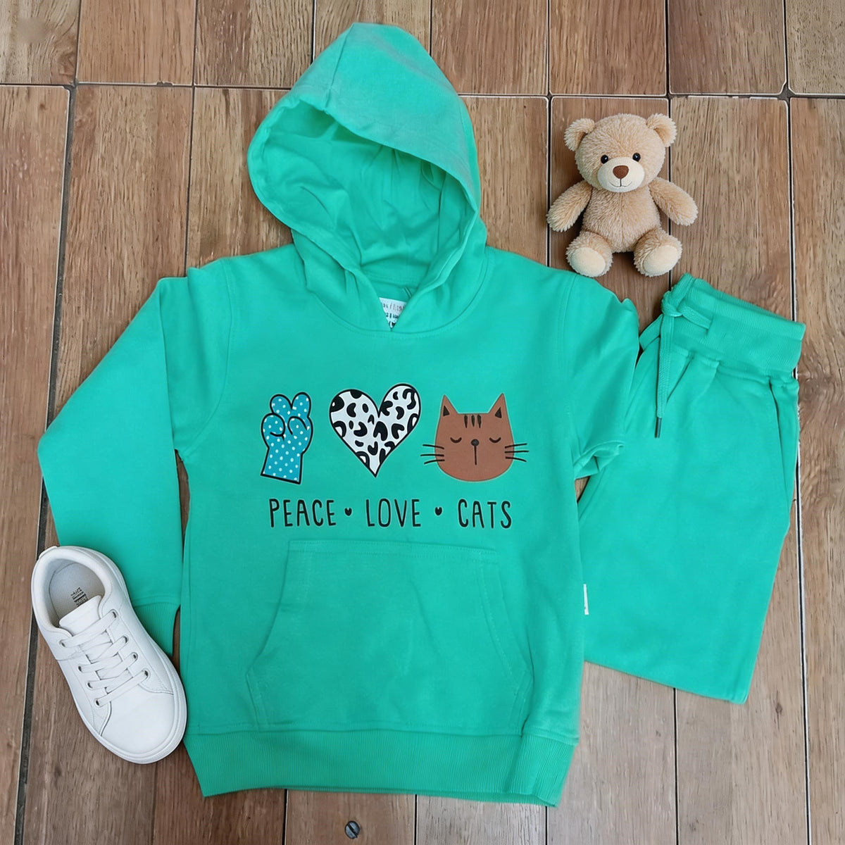 The mint Peace Love Cats cotton hoodie and matching joggers laid flat on a wooden floor beside a teddy bear and white trainers.