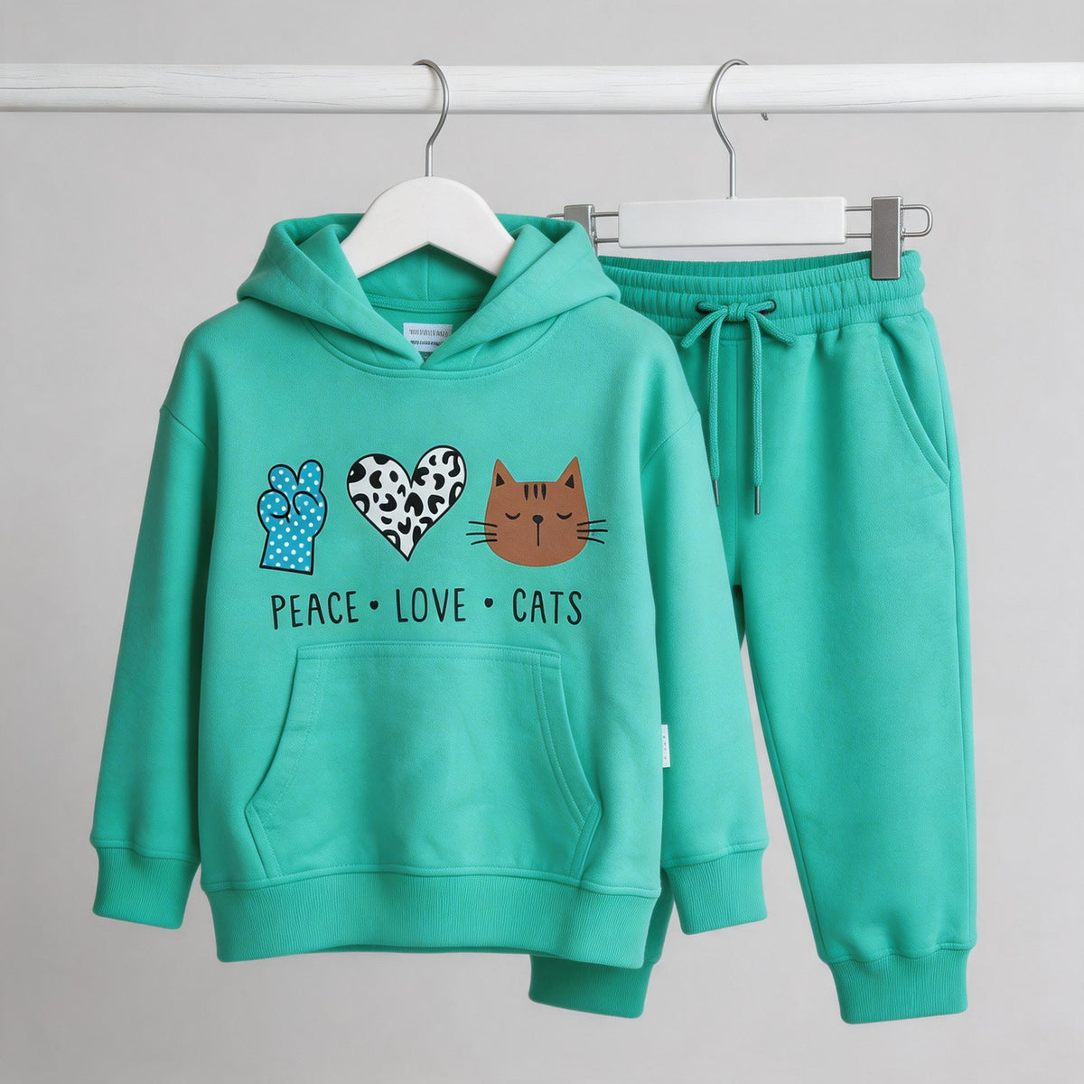 The mint Peace Love Cats cotton hoodie and matching joggers on wooden hangers against a pale wall.