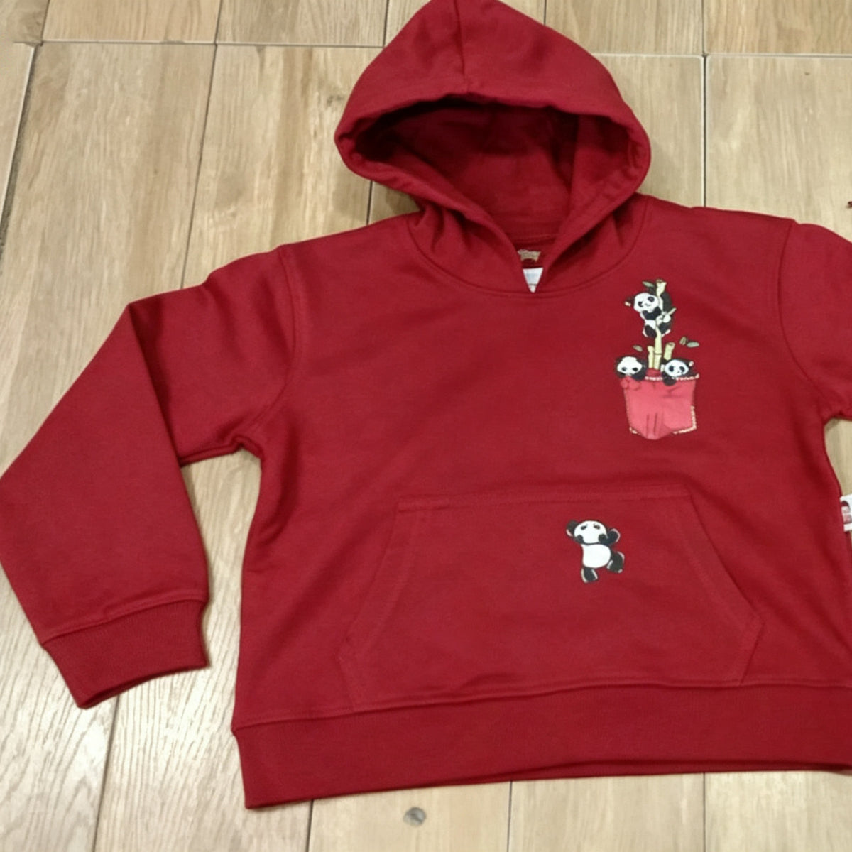 The red Panda Pocket cotton hoodie laid flat on a wooden floor, showing the printed chest pocket with pandas and the sewn front pouch.
