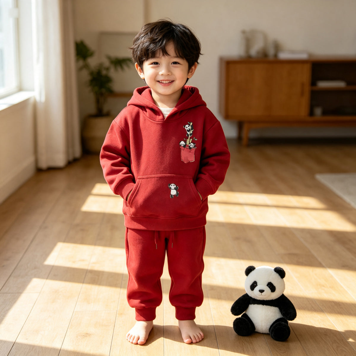 A young boy standing on a wooden floor in the red Panda Pocket cotton hoodie and jogger set, beside a toy panda. AI-generated image.