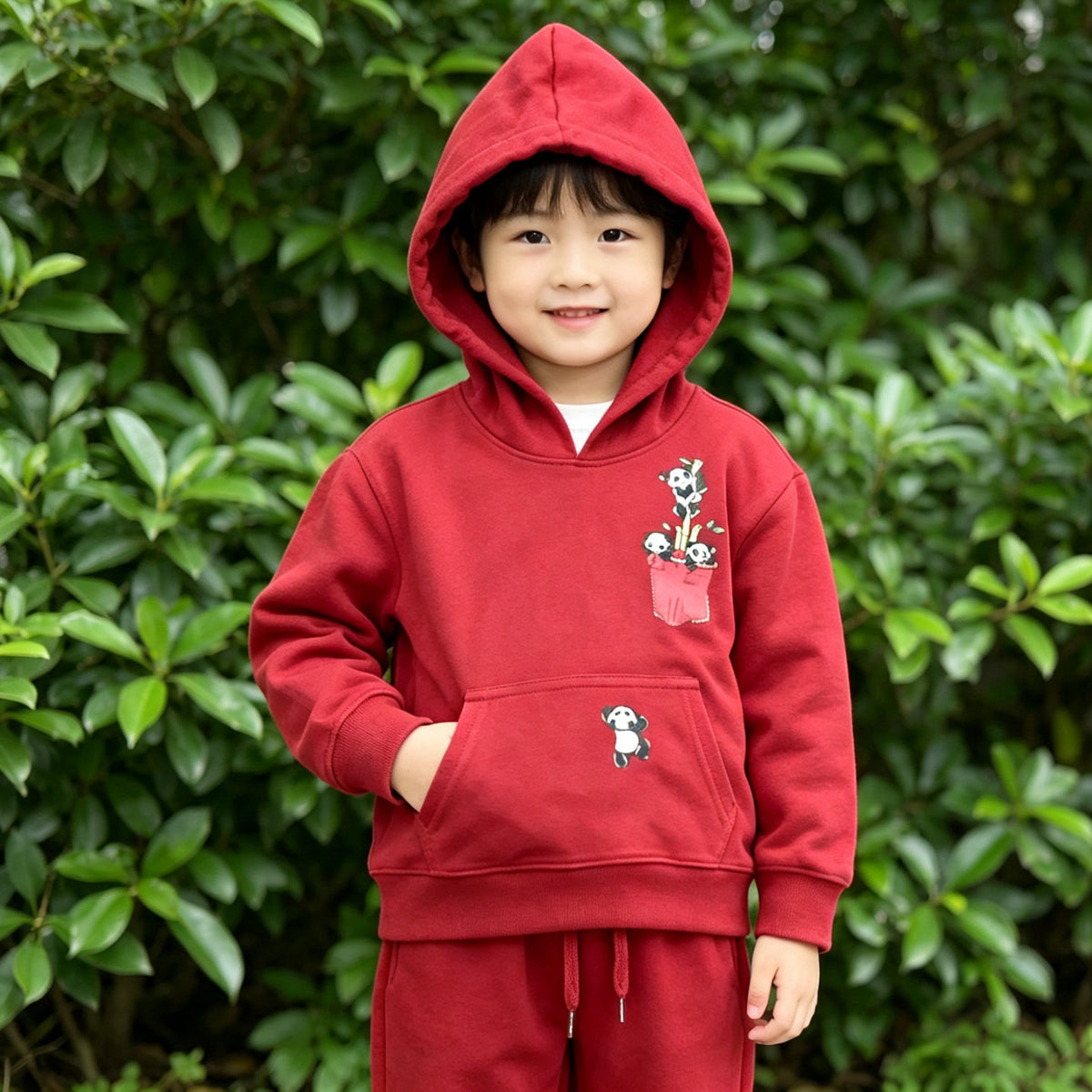 A young boy standing in front of green foliage in the red Panda Pocket cotton hoodie and jogger set, with the hood up. AI-generated image.