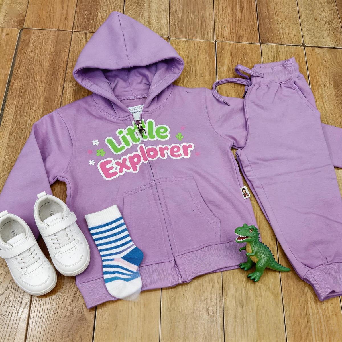 The lavender Little Explorer cotton zip hoodie and matching joggers laid flat on a wooden floor with white trainers, striped socks and a toy dinosaur.