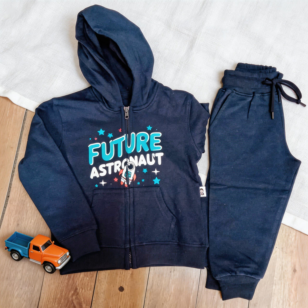 The navy Future Astronaut cotton zip hoodie and matching joggers laid flat on a wooden floor beside a toy truck.