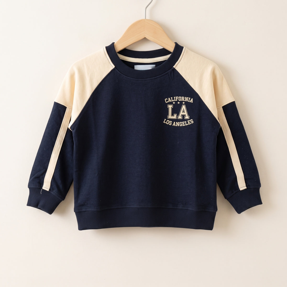 The navy cotton California LA raglan sweatshirt on a wooden hanger against a plain pale wall, showing the cream raglan yoke, the cream stripe down each sleeve, the ribbed neck, cuffs and hem, and the printed CALIFORNIA LA LOS ANGELES chest graphic.