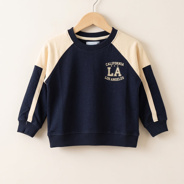 The navy cotton California LA raglan sweatshirt on a wooden hanger against a plain pale wall, showing the cream raglan yoke, the cream stripe down each sleeve, the ribbed neck, cuffs and hem, and the printed CALIFORNIA LA LOS ANGELES chest graphic.