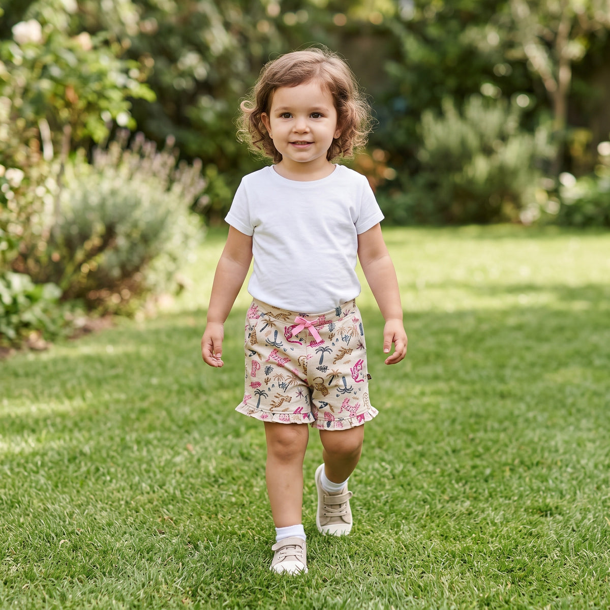 A young child walking on grass in a garden, wearing the jungle print cotton shorts with a pink ribbon bow and ruffle hems. AI-generated image.