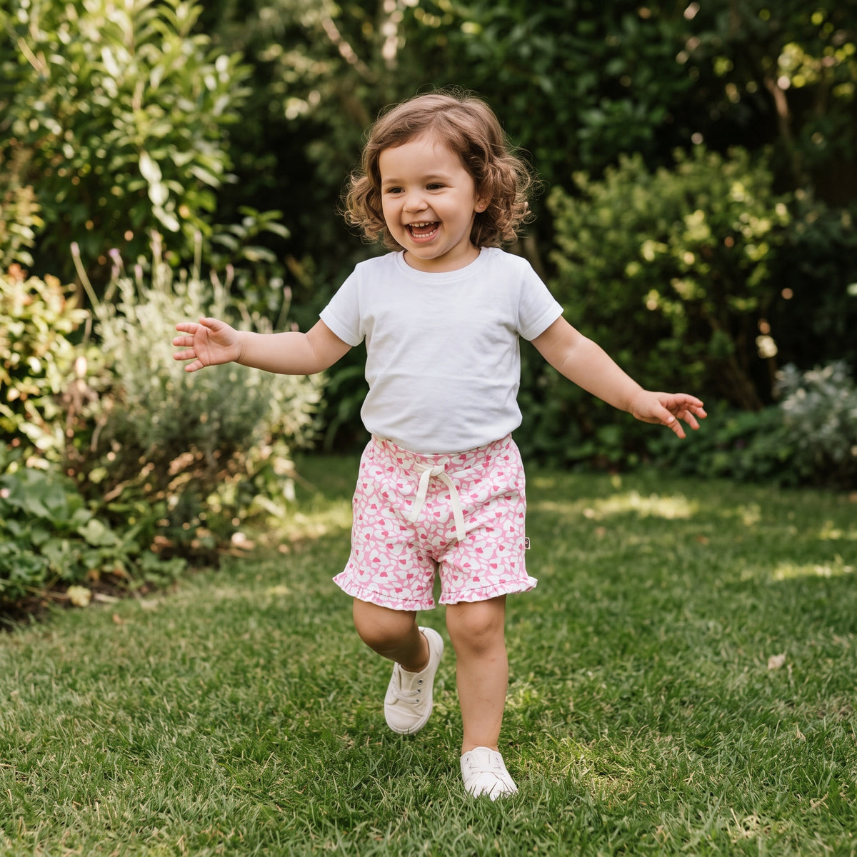 A young child running on grass in a garden, wearing the pink hearts print cotton shorts with a cream ribbon bow and ruffle hems. AI-generated image.