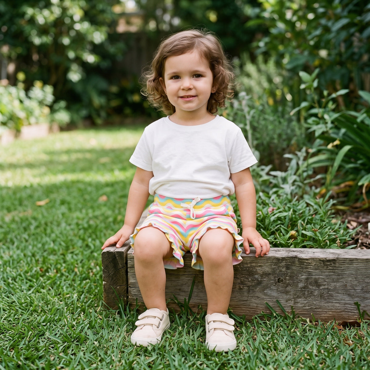 A young child sitting on a garden step, wearing the rainbow waves print cotton shorts with a cream ribbon bow and ruffle hems. AI-generated image.