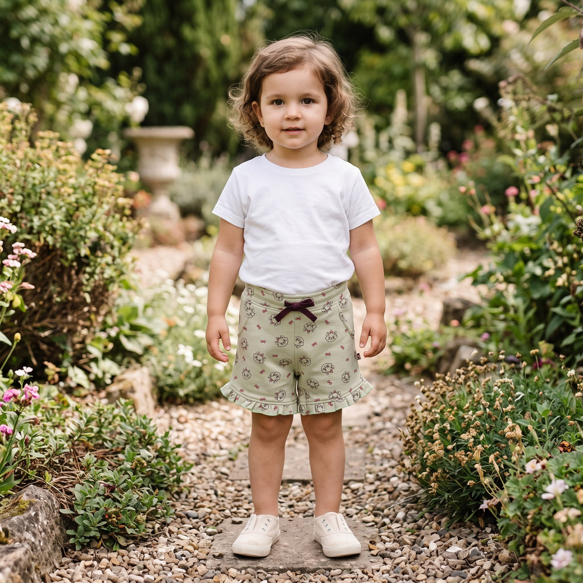 A young child standing on a garden path, wearing the sage green kitten print cotton shorts with a burgundy ribbon bow and ruffle hems. AI-generated image.
