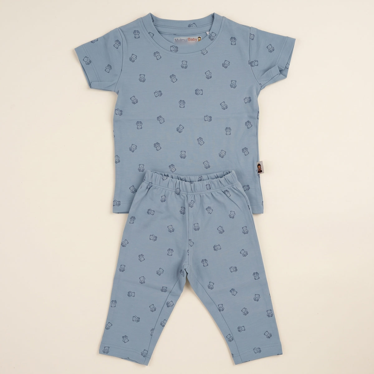 The sky blue baby bear t-shirt and matching trousers laid flat on a plain off-white ground, showing the small navy teddy bear print across both pieces, the ribbed round neck and the elastic waistband.
