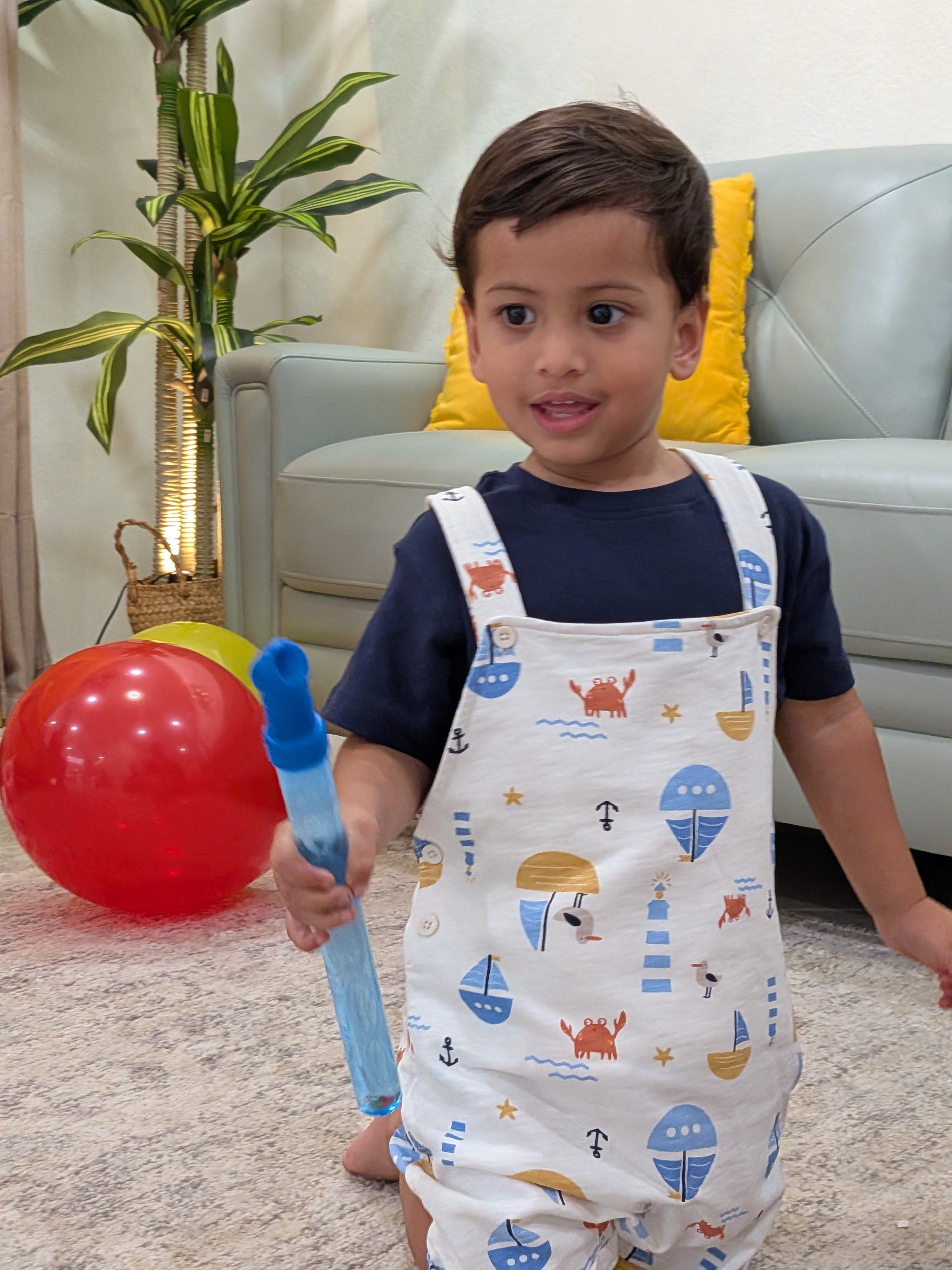 Toddler standing indoors holding a balloon wearing MulmulBaby Nautica sailboat-print organic cotton dungaree overalls set