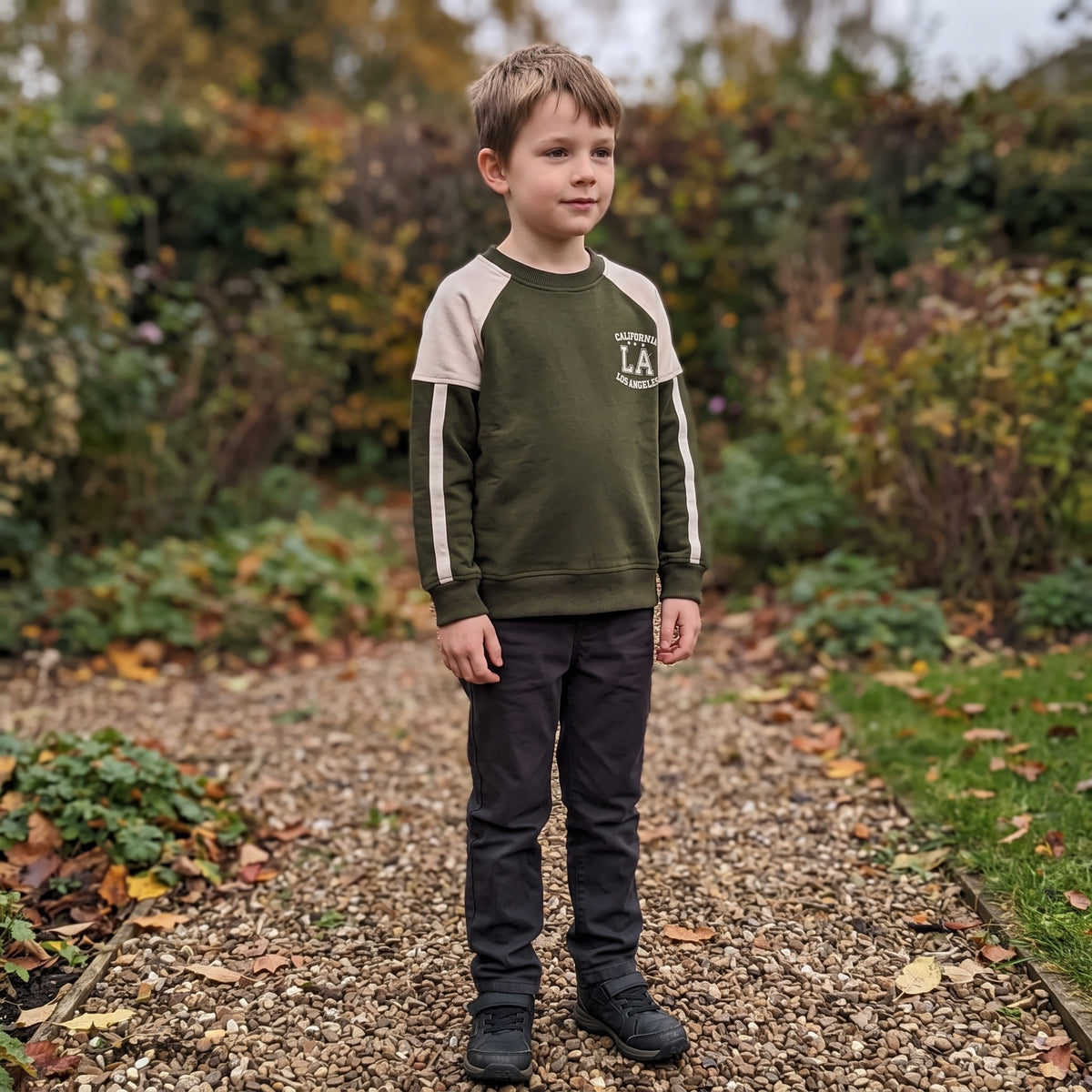 A seven-year-old standing on a garden path in autumn wearing the olive green cotton California LA raglan sweatshirt with dark trousers. AI-generated image.