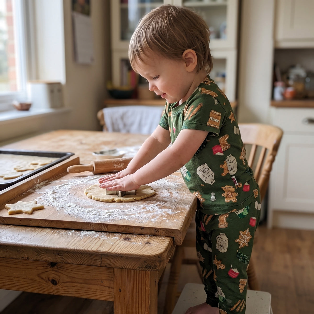 A two-year-old pressing a star cutter into rolled dough at a floured kitchen table, wearing the olive green Christmas baking print t-shirt and matching trousers. AI-generated image.