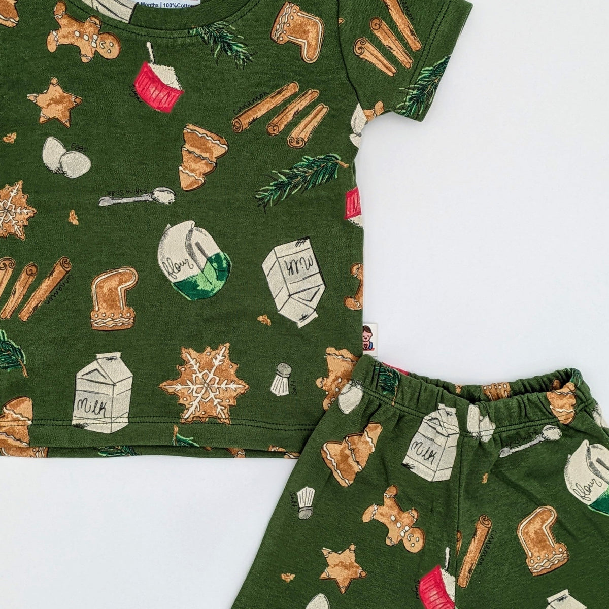 Close view of the olive green Christmas baking print showing gingerbread men, iced snowflake cookies, cinnamon sticks, milk cartons, flour bags and pine sprigs across the tee and the elastic waistband of the trousers.